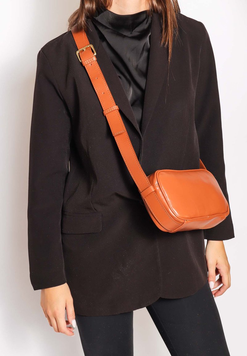 Femme portant un blazer et un pantalon noirs avec un sac bandoulière en cuir orange traversant la poitrine sur un fond blanc.