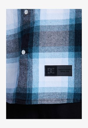 Koszula flanelowa w kratę z wzorami w kolorach niebieskim, czarnym i białym, z białymi guzikami i czarną naszywką z logo DC na dole.