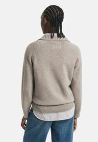 Persona con capelli intrecciati che indossa un maglione lavorato a maglia beige sopra una camicia a righe e jeans blu, vista da dietro su sfondo bianco.