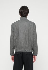 Grå, tekstureret jakke med høj krave og elastisk kant, prydet med et enkelt design. Båret over sorte bukser, set fra ryggen.