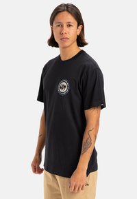 Camiseta de algodón negra con un logo gráfico redondo en el pecho, mangas cortas y un corte relajado. Presenta una textura suave y un escote redondo.