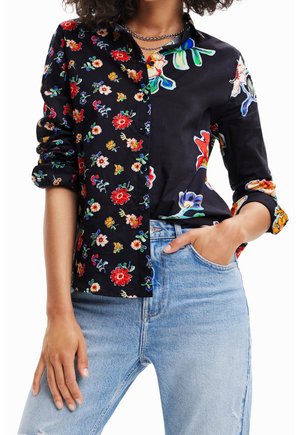 Femme portant une chemise noire à boutons avec des motifs floraux divisés et un jean bleu clair, une main dans la poche et les manches retroussées.