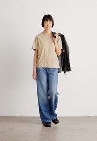 Donna che cammina indoors indossando una t-shirt beige, jeans larghi blu, scarpe nere, portando una giacca di pelle nera sopra la spalla contro un muro bianco semplice.