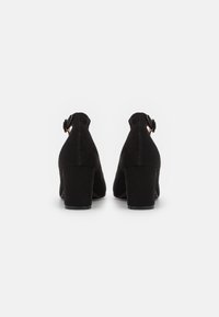 Mustad suede kontsad jalatsid, millel on paks konts ja pahkluu rihm. Tagantvaates on näha pandla detail ja sile viimistlus, mis rõhutab kinga kuju.