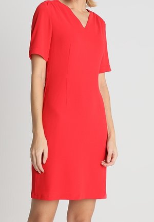 Kobieta w czerwonej sukience o długości do kolan, z krótkim rękawem i dekoltem w serek, stojąca na tle jednolitego jasnoszarego tła.