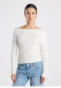 White long-sleeve top with a smocked texture, boat neckline, and frilled edges, paired with light blue jeans. Smooth, soft fabric.