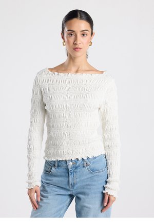 Haut blanc à manches longues avec une texture smockée, encolure bateau et bords froncés, associé à un jean bleu clair. Tissu lisse et doux.