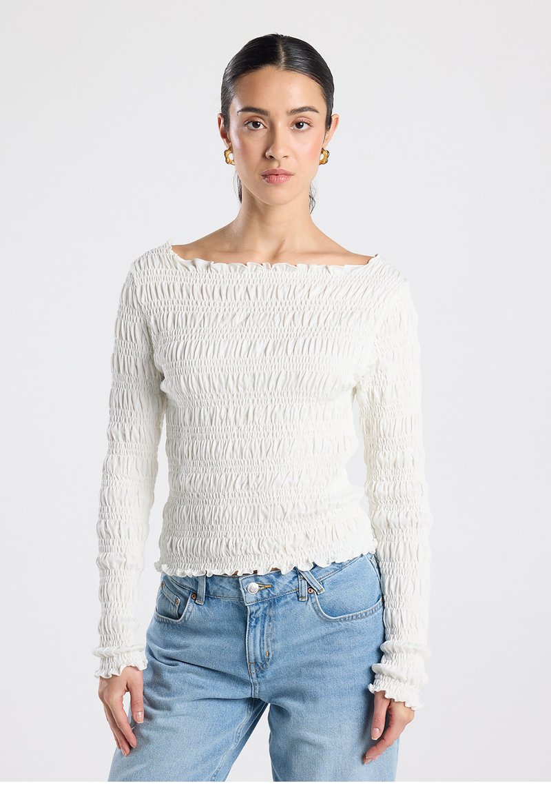 White long-sleeve top with a smocked texture, boat neckline, and frilled edges, paired with light blue jeans. Smooth, soft fabric.