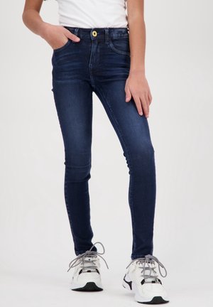 Bambino che indossa jeans skinny blu scuro con tasche anteriori e sneakers bianche chunky, in piedi con una mano in tasca contro uno sfondo semplice.