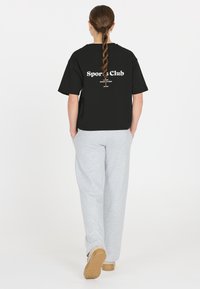 Sort t-shirt med korte ærmer med teksten "Sports Club" på ryggen, matchet med lysegrå sweatpants og beige fodtøj. Glat bomuldsstruktur.