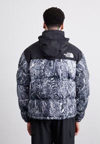 Dunjakke med sort top og abstrakt gråt og hvidt mønster, med hætte og logo på kraven. Tekstureret quiltet design.