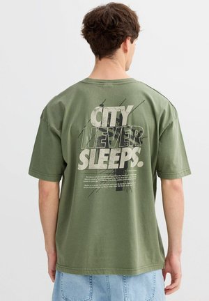 Joven con camiseta verde oliva con texto y diseño gráfico "LA CIUDAD NUNCA DUERME." en la parte trasera, combinado con vaqueros azul claro.