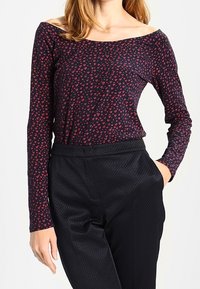 Mujer con una blusa negra de manga larga y hombros al descubierto con patrones de corazones rojos, y pantalones negros de talle alto y texturizados, con una mano en el bolsillo.