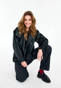 Młoda kobieta klęcząca, uśmiechnięta, ubrana w czarną, oversize'ową skórzaną kurtkę, czarne dżinsy, czerwone skarpetki i czarne loafersy na platformie.