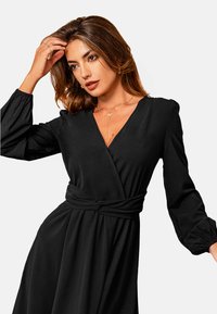 Vestido negro ajustado con mangas largas abullonadas, escote en V y cintura ceñida con cinturón. Tela suave con una textura lisa, adecuada para diversas ocasiones.