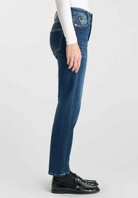 Jean en denim bleu skinny à taille haute, doté de cinq poches, d'une fermeture éclair et d'une légère décoloration le long des jambes. Porté avec des mocassins noirs.