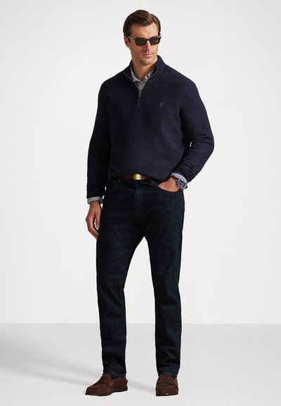 Ναυτικό ζακέτα με φερμουάρ, τζιν παντελόνια και καφέ σουέτ παπούτσια. Ορατά μανίκια ριγέ πουκαμίσου. Το μοντέλο φοράει γυαλιά ηλίου και είναι σε στάση όρθιας πόζας μπροστά σε φωτεινό φόντο.