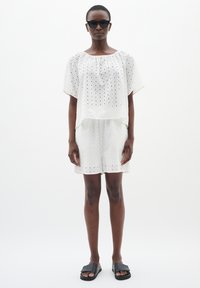 Top bianco, leggermente aderente, con un motivo testurizzato e maniche corte, abbinato a pantaloni shorts coordinati. Il modello indossa occhiali da sole neri e sandali scivolati.