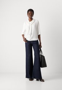 Witte blouse met een ruchesnek en een tunnelkoord, gecombineerd met een donkerdenim flared broek. Gecompleteerd met een zwarte tas en sleehak sandalen.