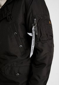 Manche de veste noire avec poche zippée et étiquette en tissu blanc comportant la mention « Remove Before Flight » attachée à la tirette du zip.