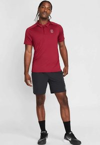 Vyras dėvi bordo spalvos trumpomis rankovėmis polo marškinėlį, juodus sportinius šortus, juodas kojines ir juodus Nike sportbačius, stovintis priešais paprastą šviesią foną.