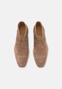 Scarpe derby in suede marrone con lacci sottili e punta affusolata. Presentano una suola di un marrone chiaro a contrasto e dettagli cuciti lungo il bordo.
