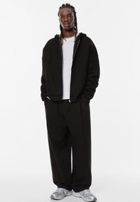 Felpa nera con cappuccio e zip argentata, abbinata a pantaloni neri a vestibilità comoda. Il modello indossa un maglione bianco a coste sotto e sneakers grigie.