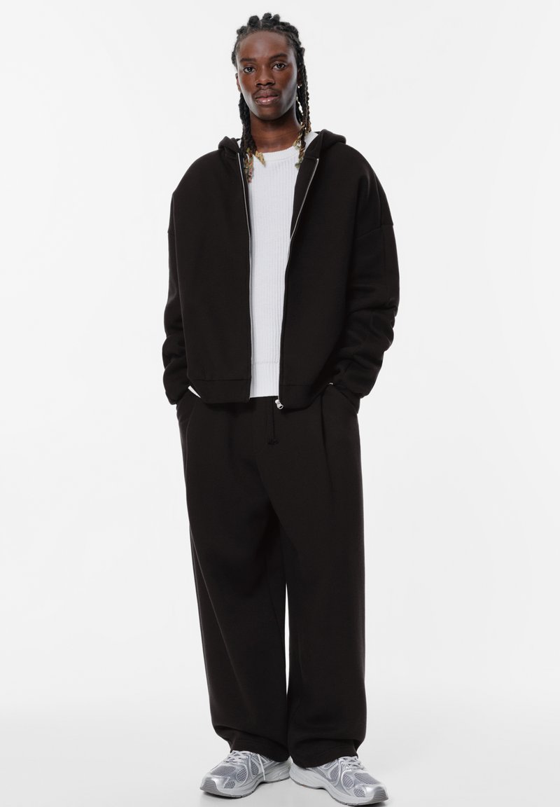 Felpa nera con cappuccio e zip argentata, abbinata a pantaloni neri a vestibilità comoda. Il modello indossa un maglione bianco a coste sotto e sneakers grigie.