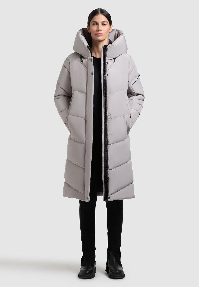 Abrigo acolchado gris claro con gran capucha, bolsillos frontales y cierre de cremallera. Presenta un diseño acolchado con líneas horizontales y un acabado ligeramente brillante.