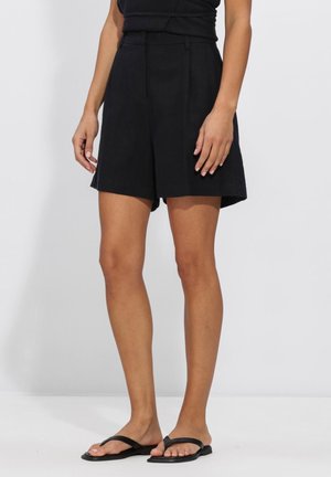 Vrouw draagt zwarte, hooggetailleerde plisséshorts en zwarte platte teenslippers, staand tegen een effen lichte achtergrond.