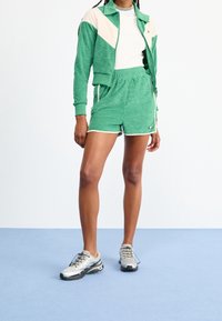 Grön velourjacka med krämfärgad chevron, tillsammans med gröna shorts. Matchningen kompletteras av grå och svarta sportskor.