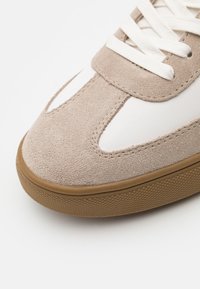 Baskets en daim beige clair avec accents en cuir blanc, semelle en caoutchouc texturée et lacets plats. Présente un bout arrondi et des détails de couture subtils.