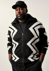 Schwarz-weißer, gezackter Strick-Zip-up-Pullover mit hohem Kragen, langen Ärmeln und weicher Textur. Über einem schwarzen Hemd getragen.