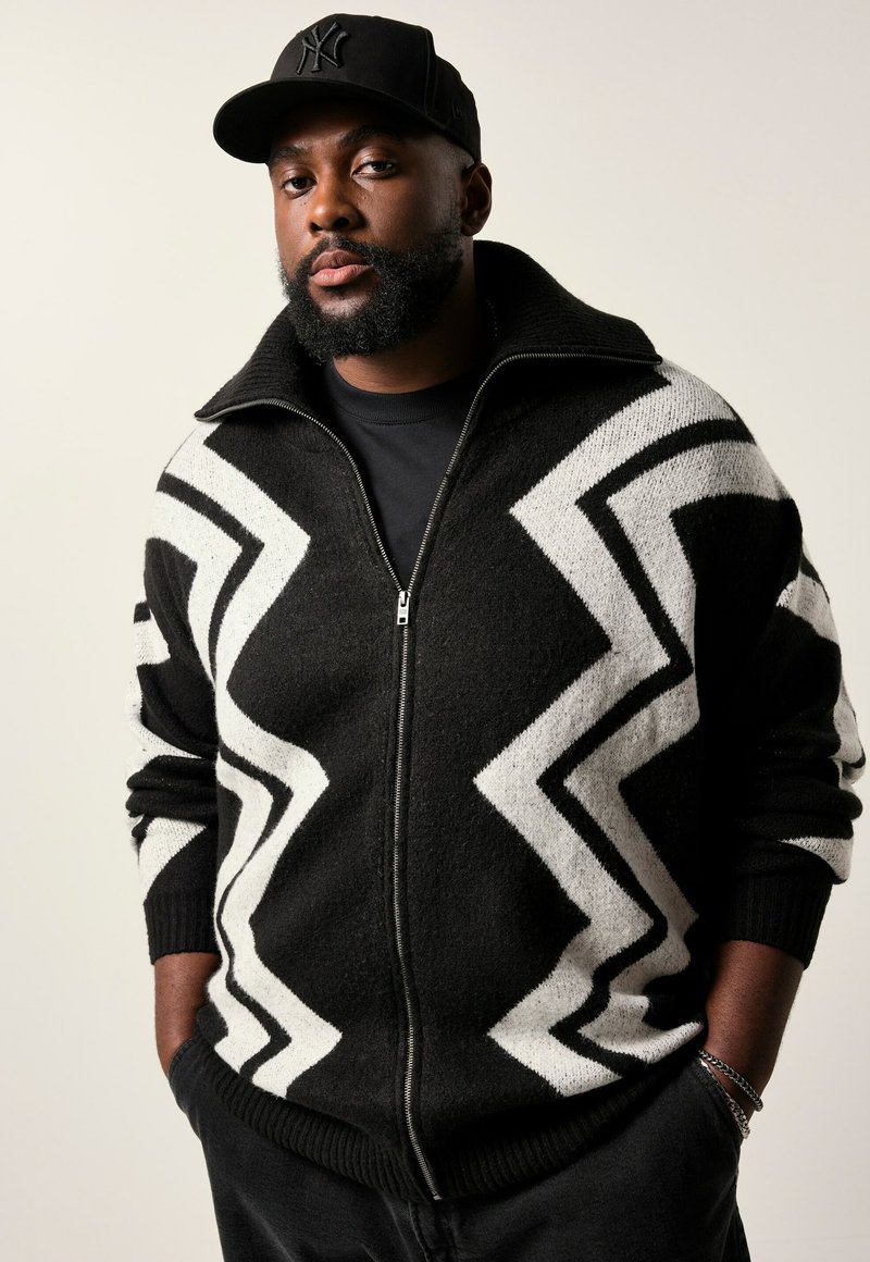 Schwarz-weißer, gezackter Strick-Zip-up-Pullover mit hohem Kragen, langen Ärmeln und weicher Textur. Über einem schwarzen Hemd getragen.