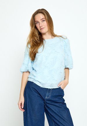 Žena s dugom svijetlosmeđom kosom nosi svijetloplavu teksturiranu bluzu i tamnoplave široke hlače ispred bijele pozadine.