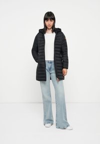 Zwarte gewatteerde jas met een capuchon, voorzien van een rits aan de voorkant en verticale stiksels, gecombineerd met lichtblauwe flared jeans en witte sneakers.