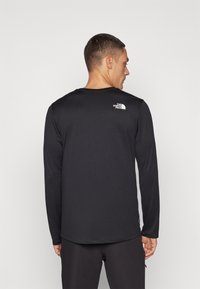 Langærmet sort skjorte lavet af letvægtsstof med et hvidt logo på den øverste del af ryggen. Glat tekstur, rund hals og syede kanter.