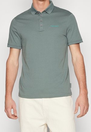 Mężczyzna w krótkim rękawie zielonej koszulce polo z trzema guzikami i białych spodniach, stojący na jednolitym tle.