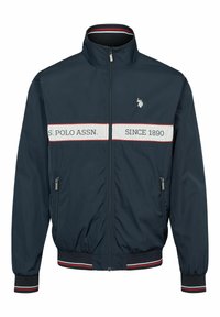 Granatowa kurtka z wysokim kołnierzem, ozdobiona białym paskowanym wzorem z napisem "S. POLO ASSN." oraz "SINCE 1890"; dwie boczne kieszenie na zamek.