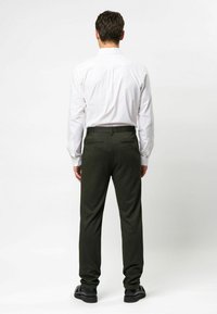 Tamsiai žalios kelnės, pagamintos iš tekstūruoto audinio, tiesaus kirpimo, su dviem užpakalinėmis kišenių, dėvimos su balta marškinėliais. Matomos juodos avalynės.