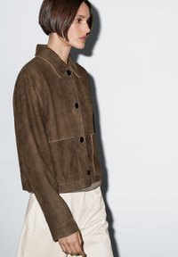 Hnědá semišová bunda s velkými černými knoflíky, špičatým límcem a dvěma předními kapsami. Design je s krátkou délkou a neutrálními tóny.