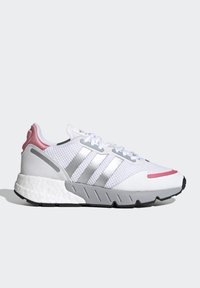 Zapatilla deportiva blanca con parte superior de malla, franjas plateadas, acento en el talón rosa y suela texturizada gris con una forma distintiva.
