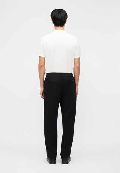 Άνδρας που φοράει απλό λευκό κοντομάνικο πουκάμισο, μαύρο παντελόνι με πίσω τσέπες και μαύρα παπούτσια, στεκόμενος με την πλάτη γυρισμένη σε λευκό φόντο.