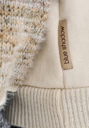 Sweter dziany wykonany z kremowej, teksturowanej tkaniny z miękkim, puszystym brzegiem oraz brązową metką z napisem „blue shadow” przymocowaną z boku.