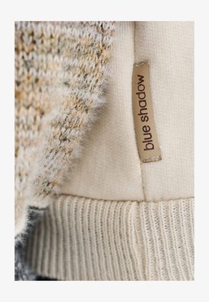Sweter dziany wykonany z kremowej, teksturowanej tkaniny z miękkim, puszystym brzegiem oraz brązową metką z napisem „blue shadow” przymocowaną z boku.