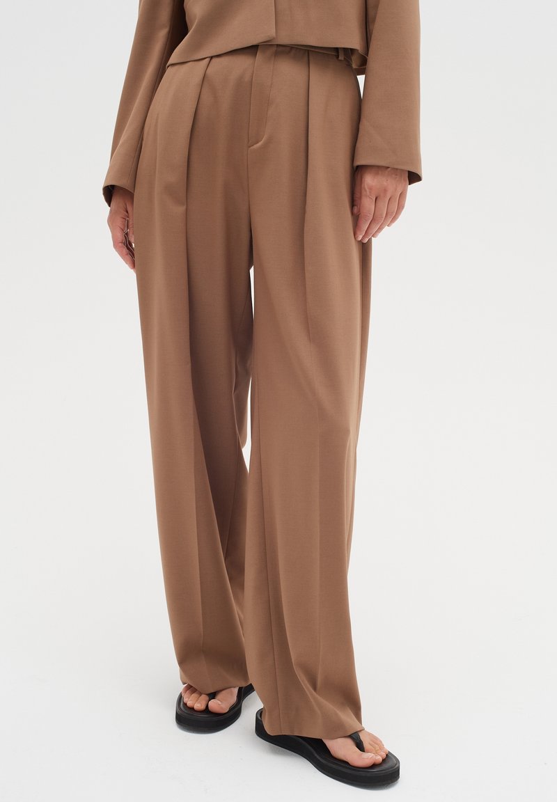 Pantalon marron à jambes larges, taille haute avec plis avant, porté avec un haut assorti marron et des sandales à plateforme noires.