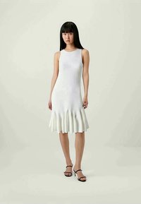 Vestido branco sem mangas com um corpete ajustado e bainha evasê. Tecido liso e texturizado. Modelo usa saltos pretos com tiras. Fundo neutro.