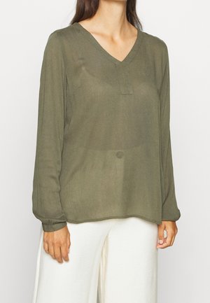 Kobieta ubrana w półprzezroczystą oliwkowozieloną bluzkę z długim rękawem i dekoltem w serek oraz białe, szerokie spodnie, stojąca na jednolitym tle.