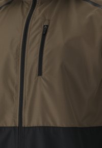 Leichte Jacke in Braun und Schwarz mit einer Reißverschlusstasche, glatter Textur und einem zweifarbigen Design mit subtiler Rautenmusterung.