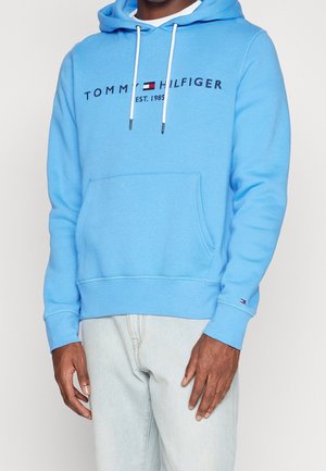 Mann trägt eine hellblaue Tommy Hilfiger Kapuzenjacke mit Fronttasche und weißen Kordeln, kombiniert mit hellfarbenen Jeans.